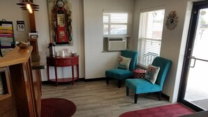 A room with a wooden reception desk, a red side table, two teal chairs, a red and white patterned rug, a wall clock, and a window with blinds.