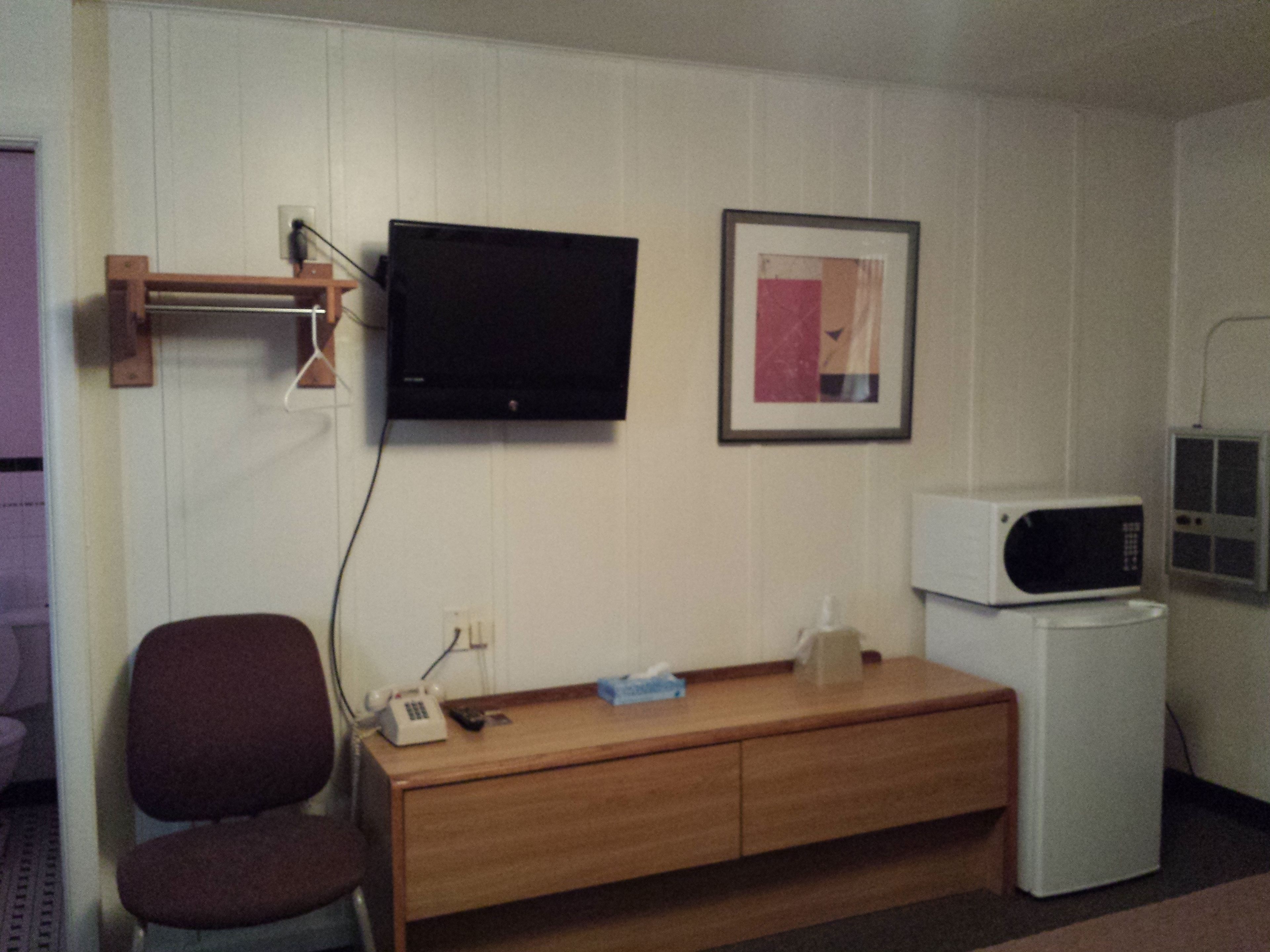 Una habitación pequeña con un escritorio de madera, una silla, un televisor montado en la pared, un microondas y un cuadro enmarcado.