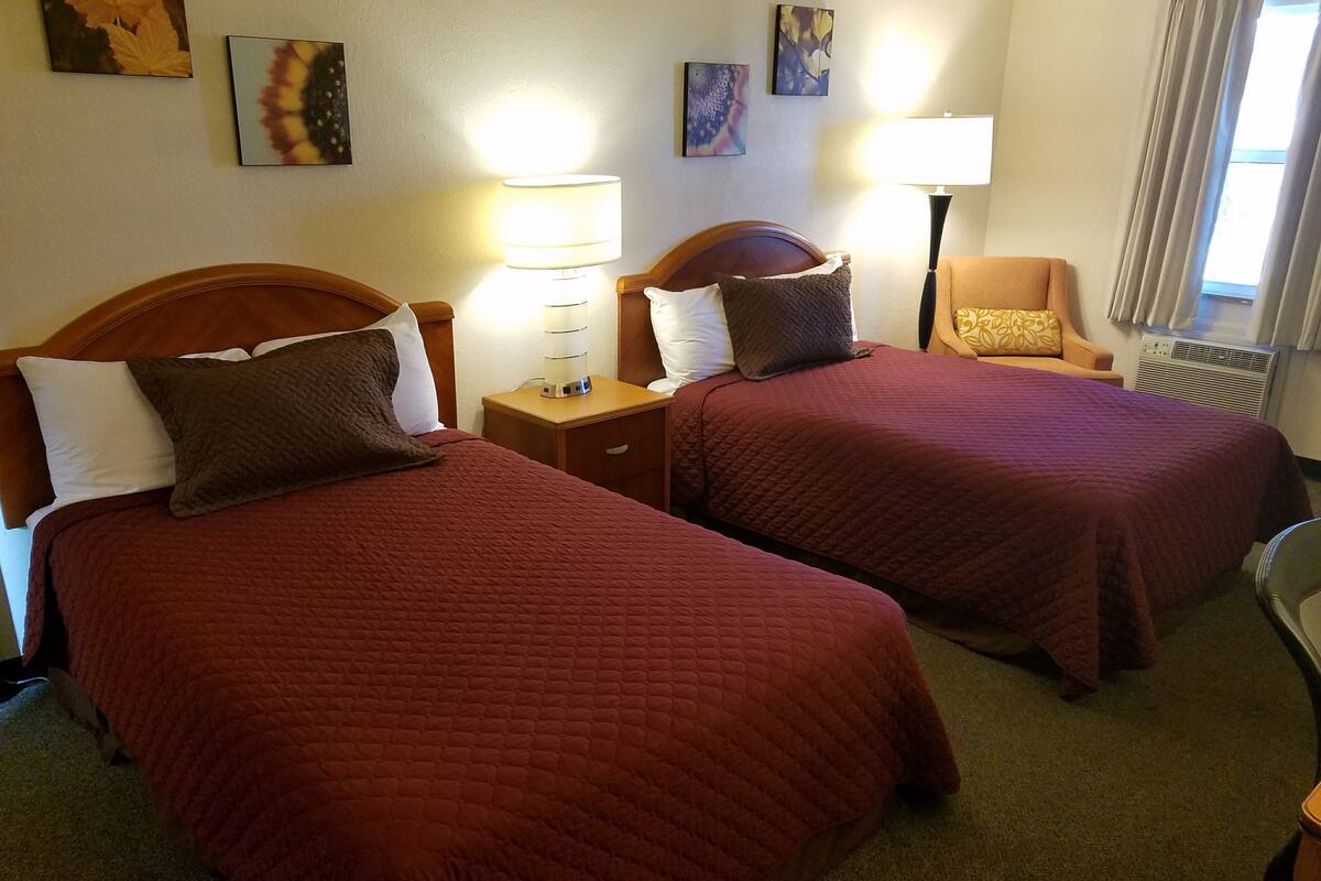Ein Hotelzimmer mit zwei Betten, jeweils mit einem hölzernen Kopfteil, einem Nachttisch mit Lampe und einem Sessel.