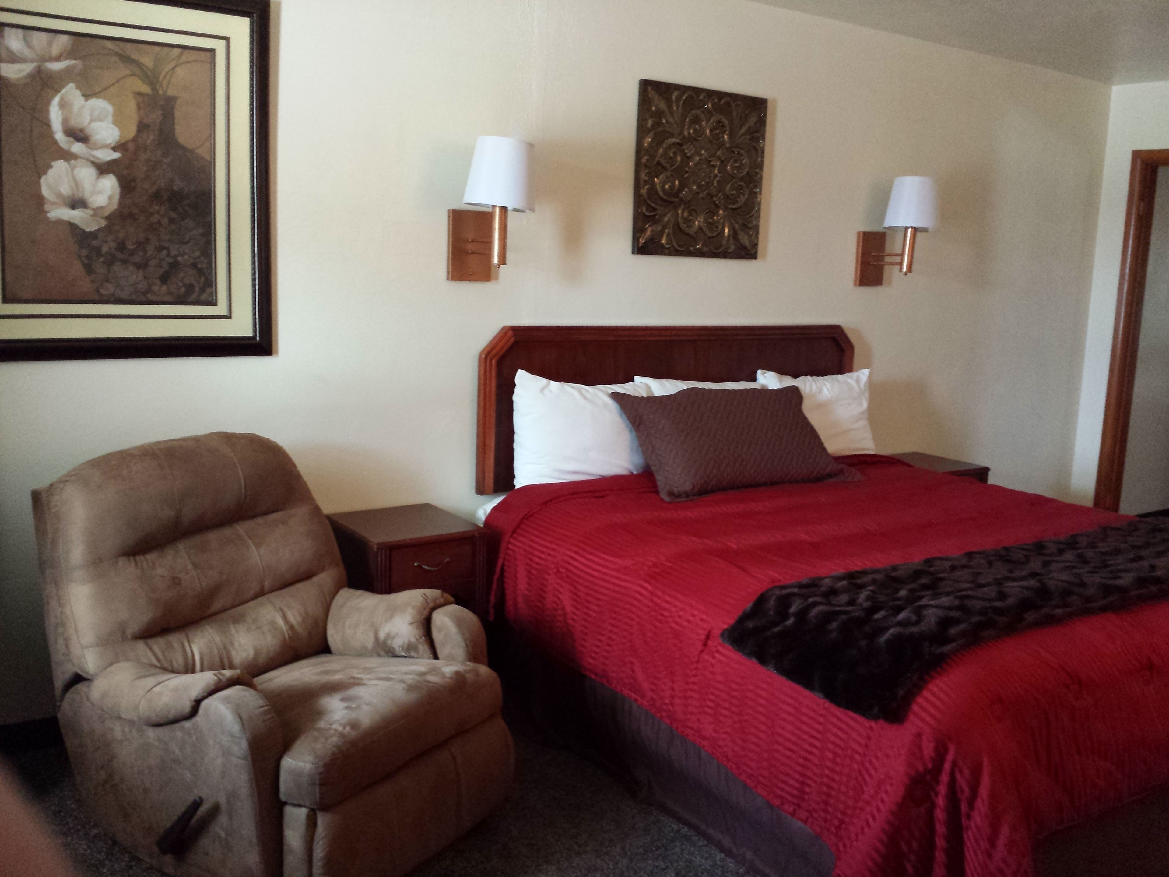 Habitación de hotel con cabecera de madera, colcha roja, sillón de cuero marrón y dos lámparas fijadas a la pared.