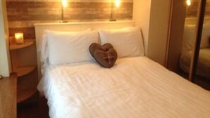 Una cama bien hecha con sábanas blancas y una almohada en forma de corazón, un cabecero de madera y dos lámparas de noche.