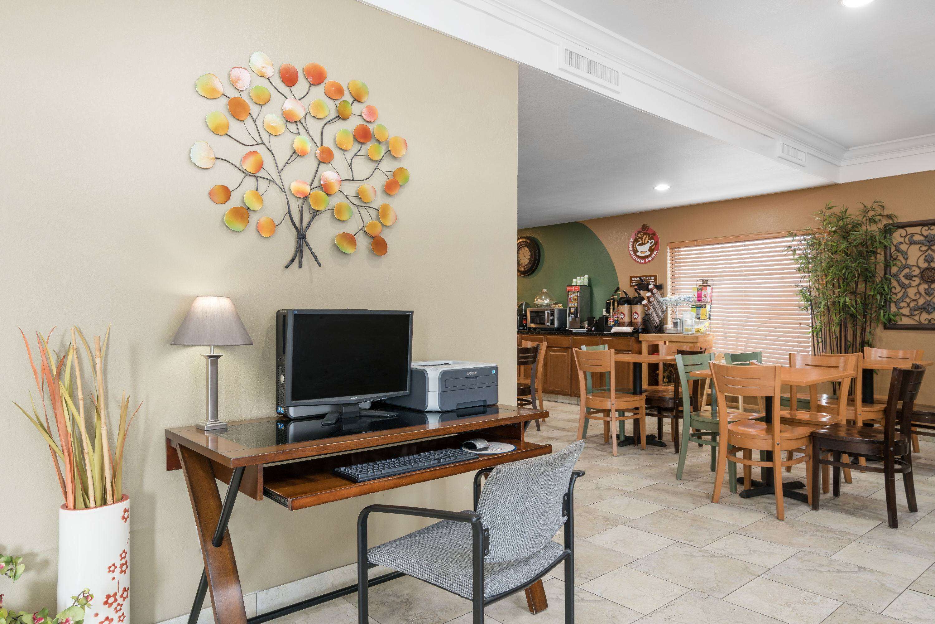 A home office with a computer, printer, and desk lamp. There is a dining area with wooden chairs and a table. A decorative tree wall art is visible.