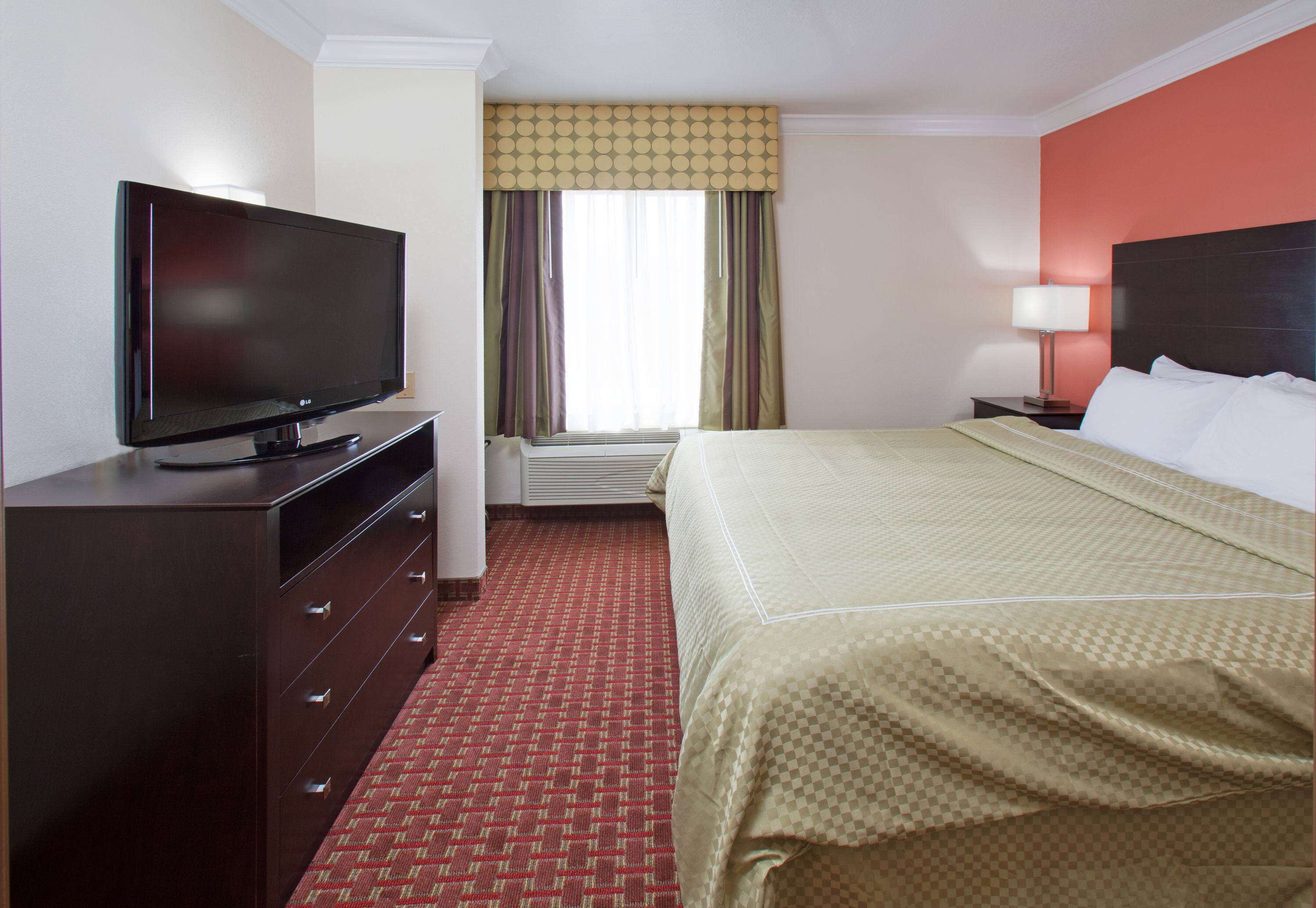 Una habitación de hotel con una cama, un televisor sobre una cómoda, una mesita de noche con lámpara y una ventana con cortinas.