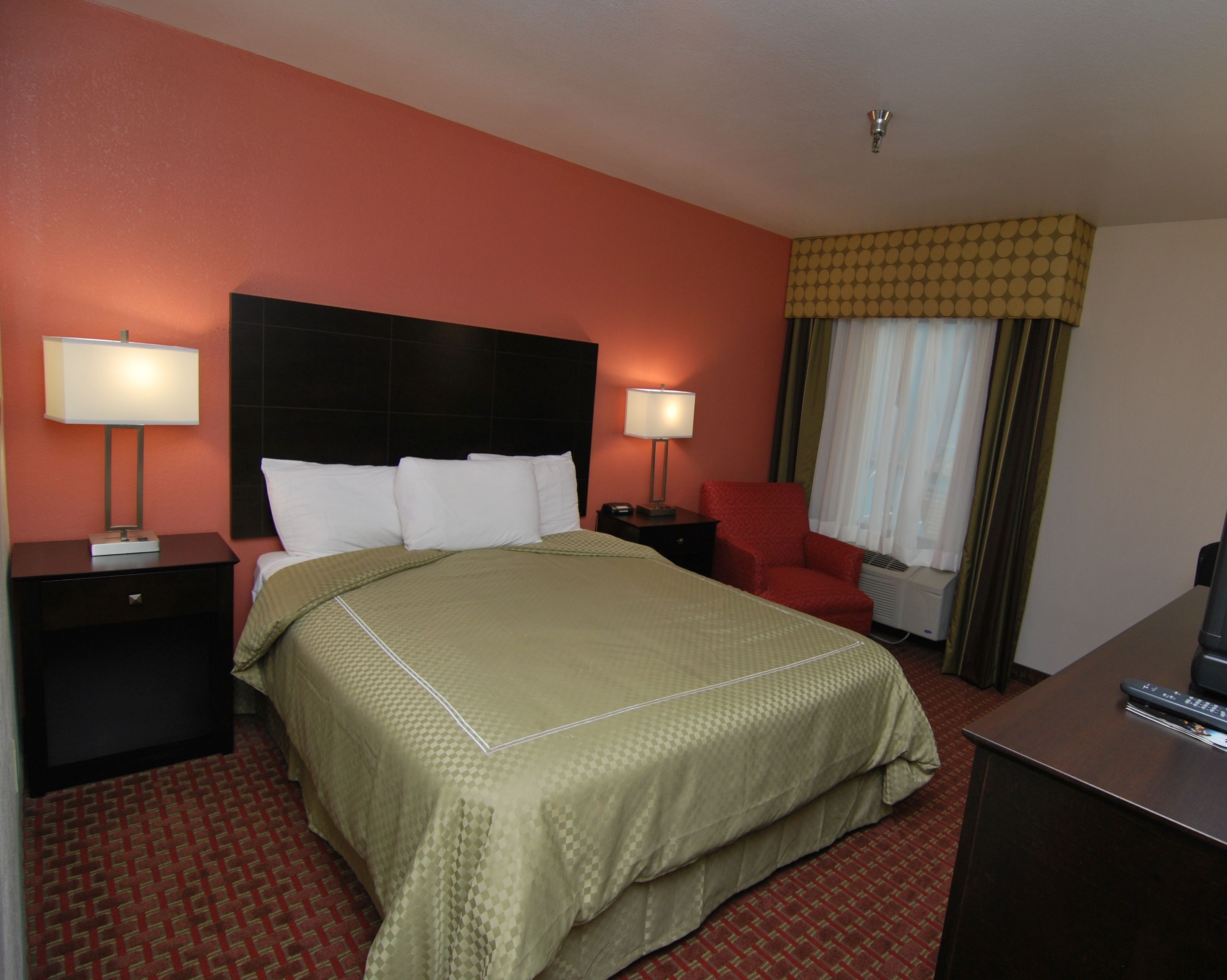 A hotel room with a large bed, two bedside tables with lamps, a red armchair, a desk with a remote control, and a window with curtains.
