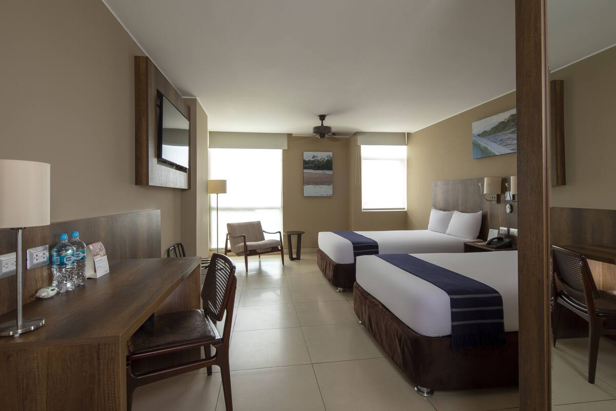 A hotel room with two beds, a desk with a chair, a small table, and a wall-mounted TV.