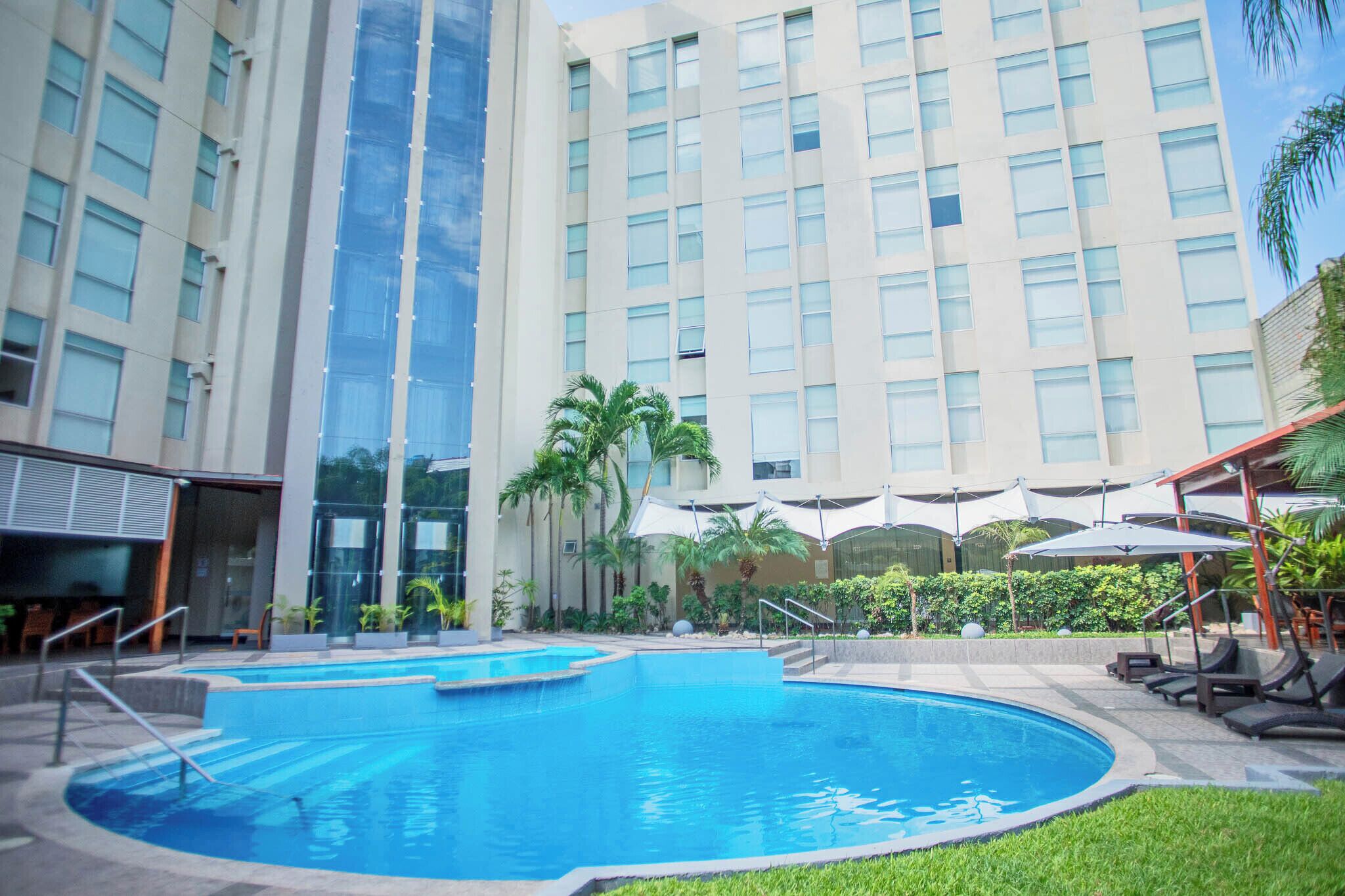 Un hotel con una amplia piscina cubierta, palmeras y una fachada de edificio moderna.