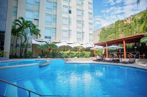 A hotel pool area with a clear blue pool, lounge chairs, umbrellas, and a building with a modern design and greenery.