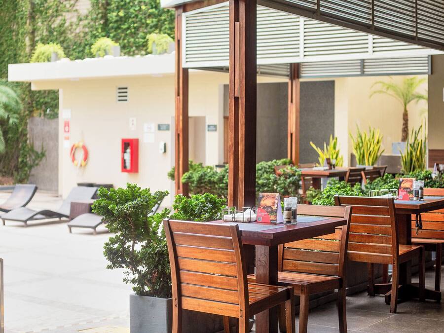 An outdoor dining area with wooden tables and chairs, surrounded by potted plants and a white building with a red door.