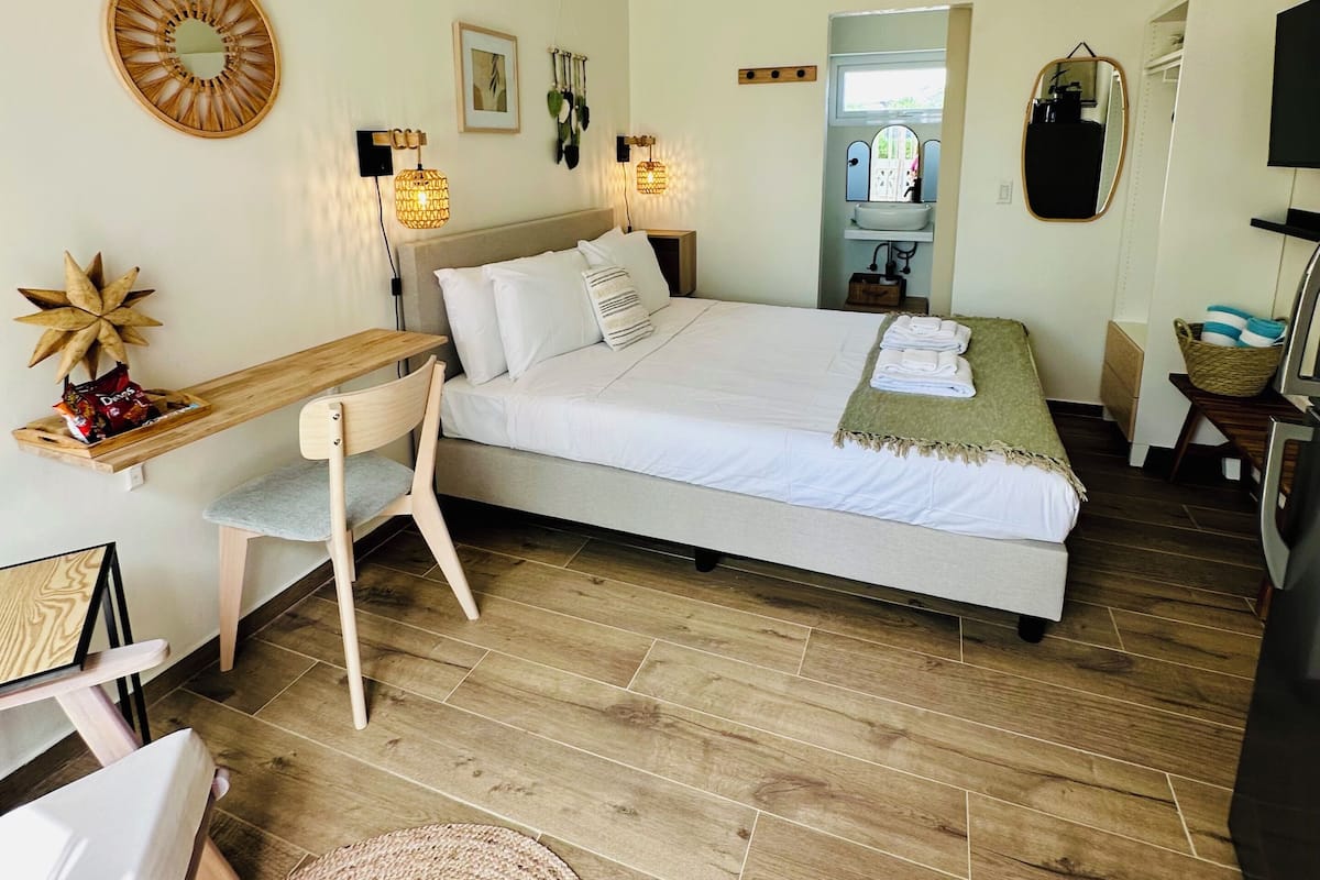 A bedroom with a bed, bedside table, chair, and a desk. There is a mirror, wall art, and an air conditioning unit.
