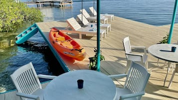 Eine Holzterrasse mit weißen Stühlen und Tischen, einem Kajak sowie einem Steg mit Booten im Hintergrund.