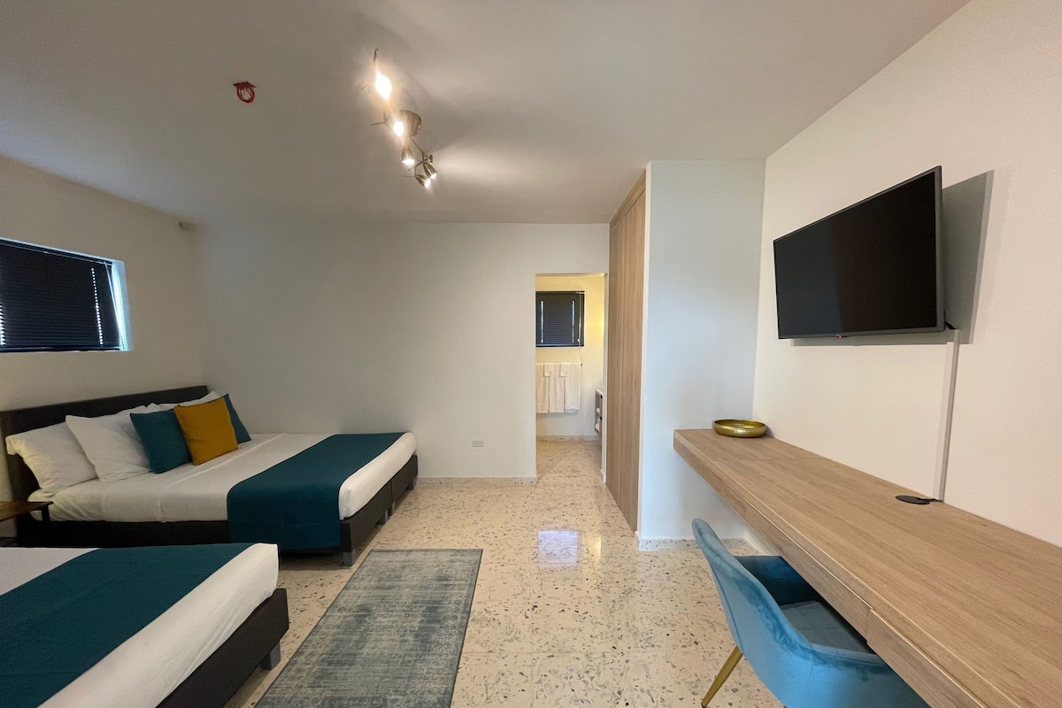A hotel room with two beds, a desk, a chair, a TV, and a bathroom visible through an open door.
