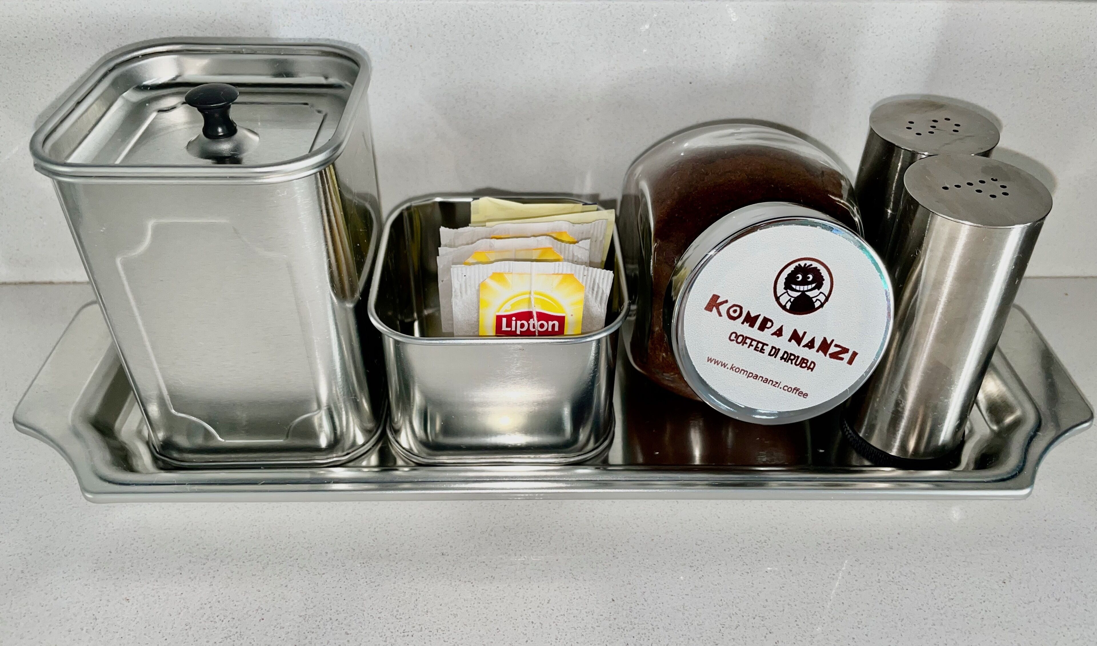 A stainless steel tray with a Lipton tea bag, a Kompananzi Coffee di Aruba tin, and two salt shakers.