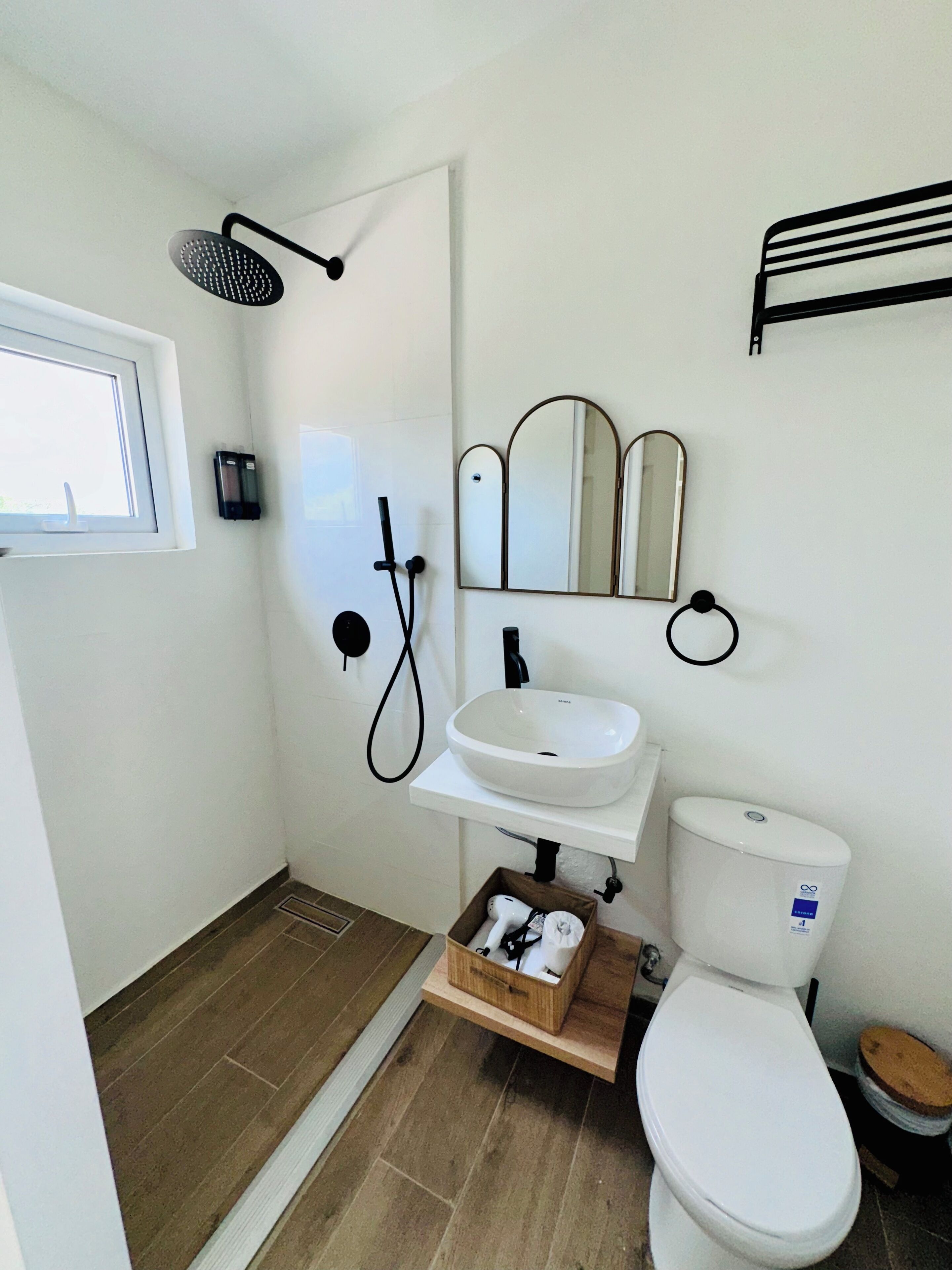 Un baño pequeño con un lavamanos blanco, un inodoro, una ducha con cabezal de ducha manual y una ventana pequeña.