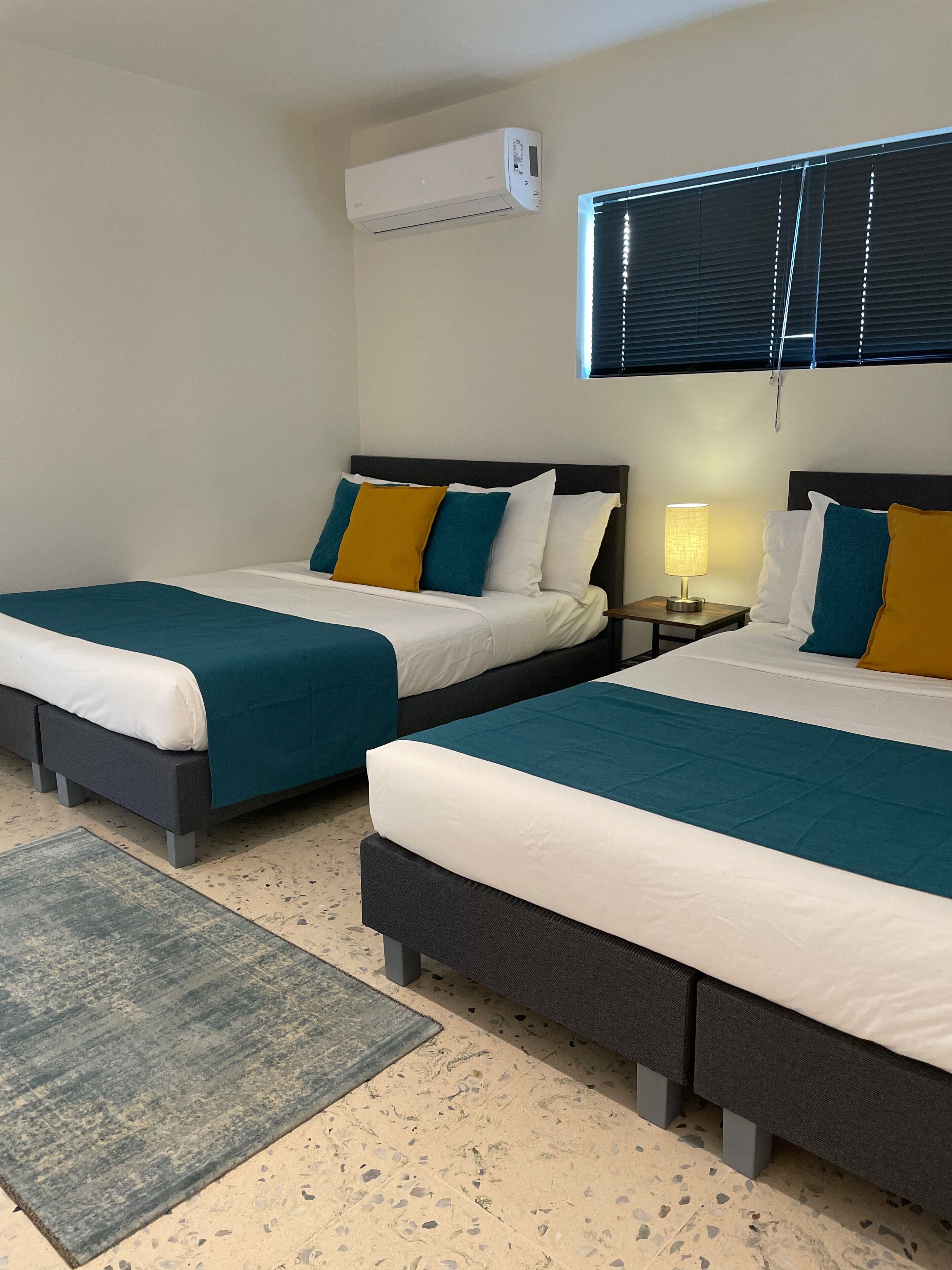 Habitación de hotel con dos camas, una mesita de noche con lámpara, un equipo de aire acondicionado y una ventana con persianas.