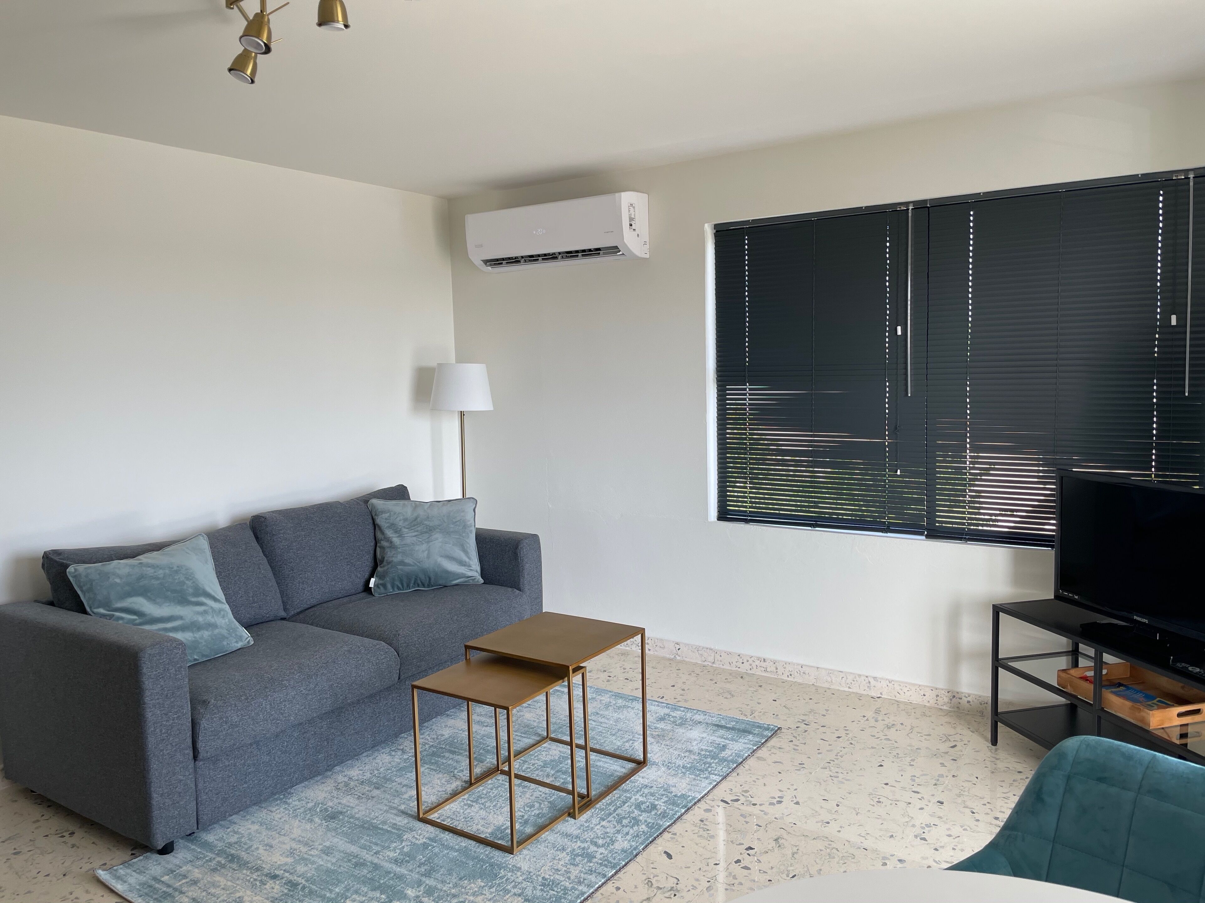 Una sala moderna con un sofá gris, una mesita de madera, un televisor sobre un mueble negro y una ventana con persianas.