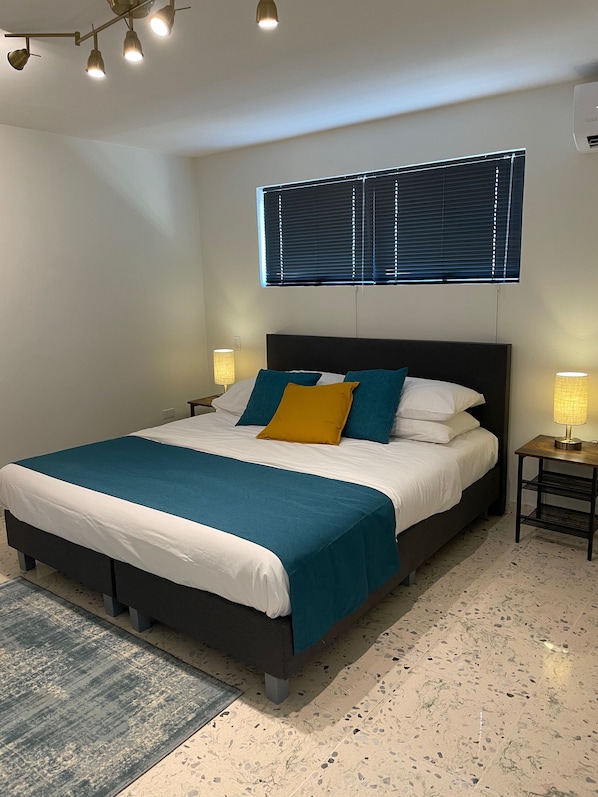 Un dormitorio ordenado con cama, mesitas de noche y una ventana con persianas.