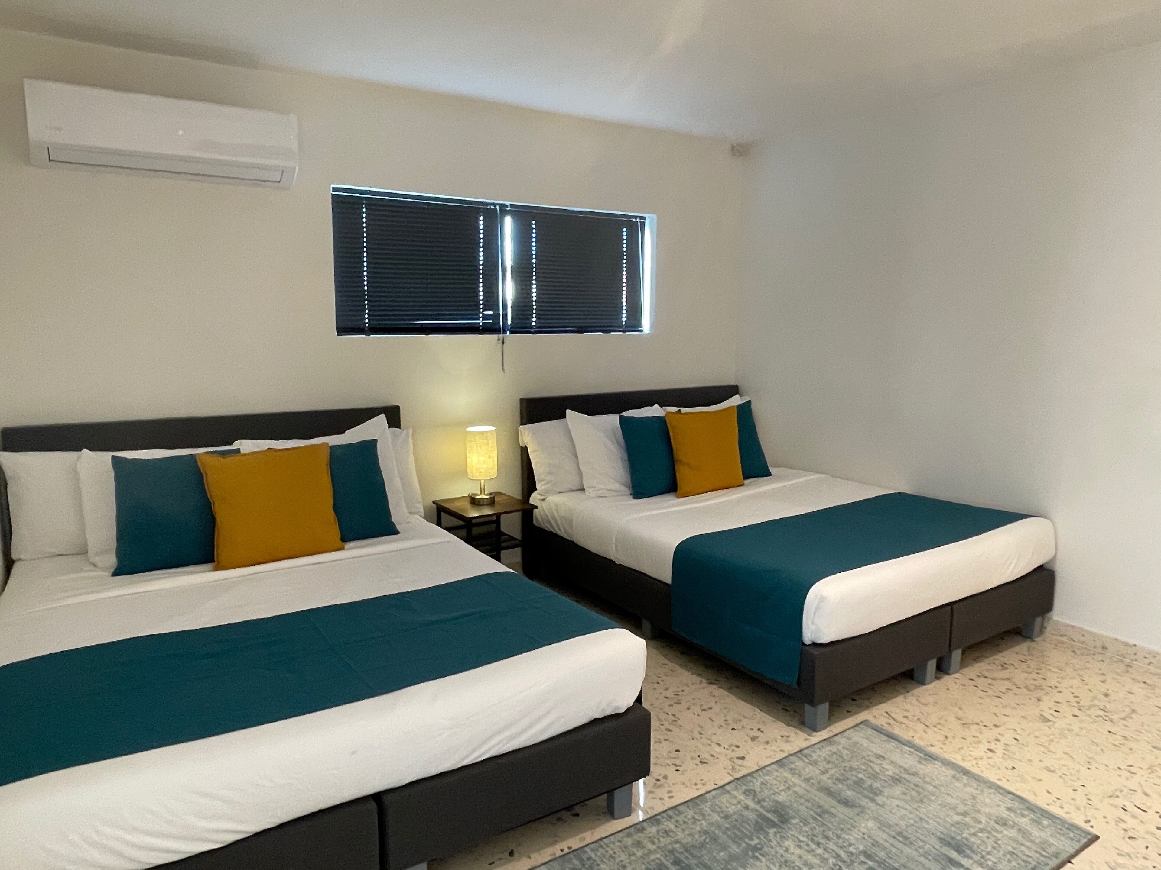 Habitación de hotel con dos camas, cada una con cubrecama azul verdoso y blanco, y una mesita de noche pequeña con lámpara.