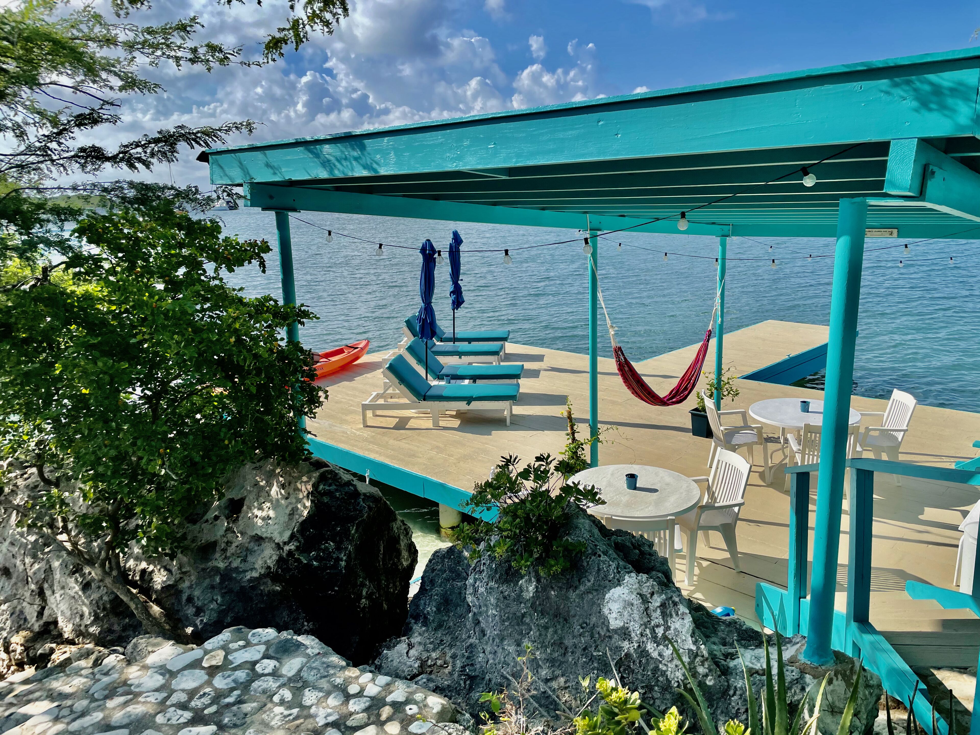 Un área de estar al aire libre color turquesa con sillones reclinables y una hamaca, con vista a un cuerpo de agua.