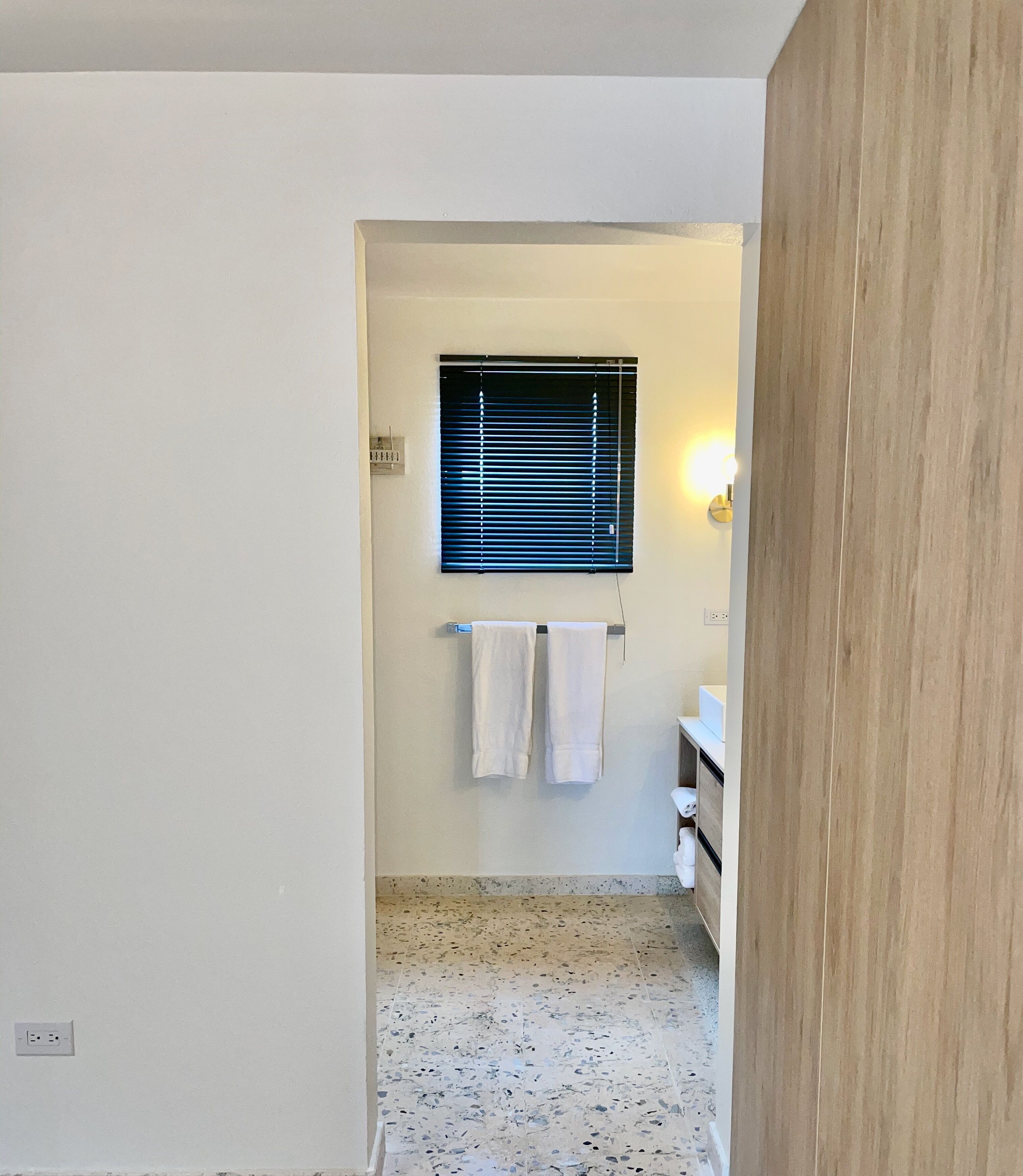 Un couloir étroit mène à une salle de bain équipée d’une fenêtre, d’un porte-serviettes et d’un lavabo.