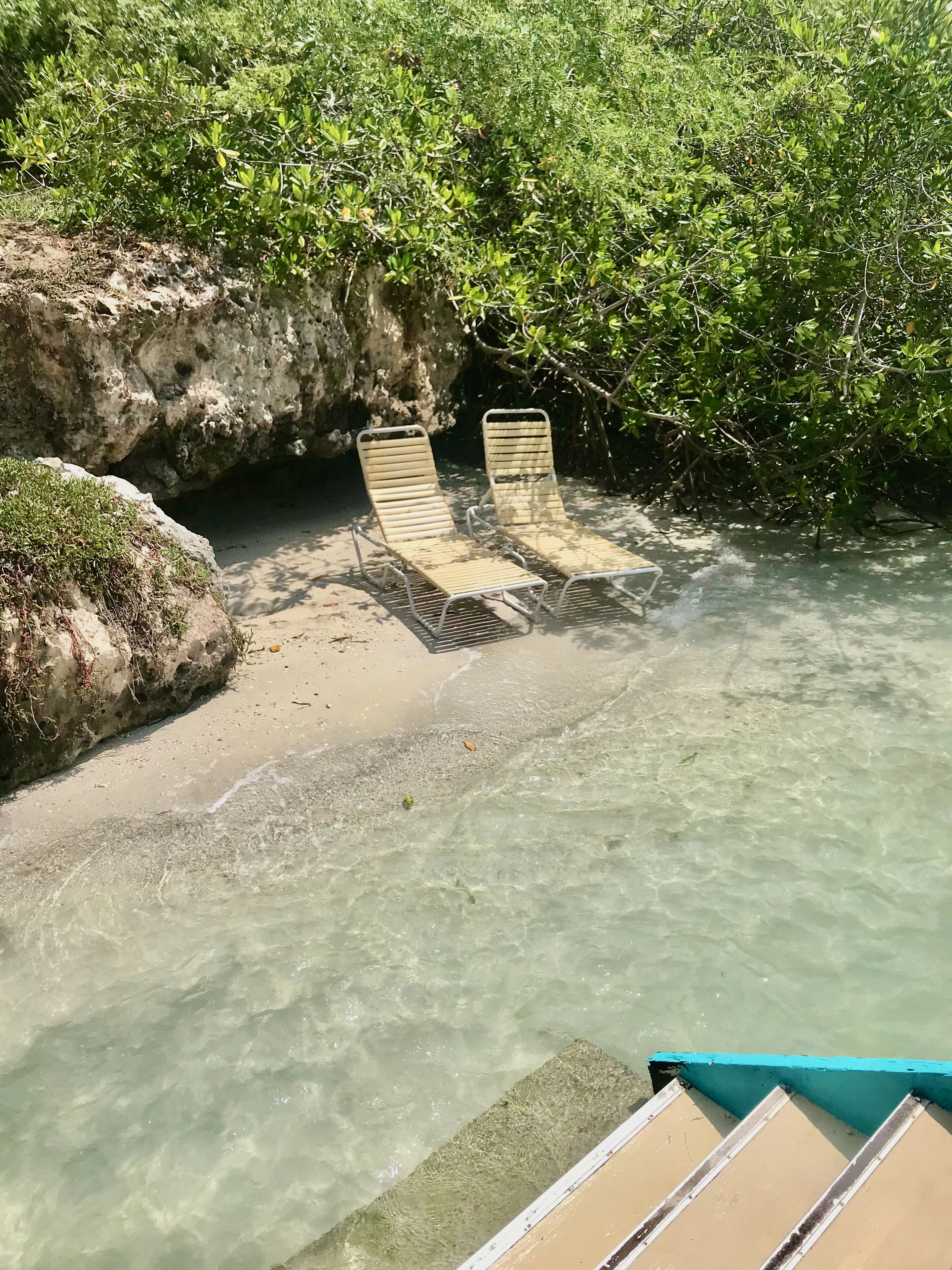 Dos sillones vacíos al lado de una piscina cristalina, con un paisaje rocoso y abundante vegetación.