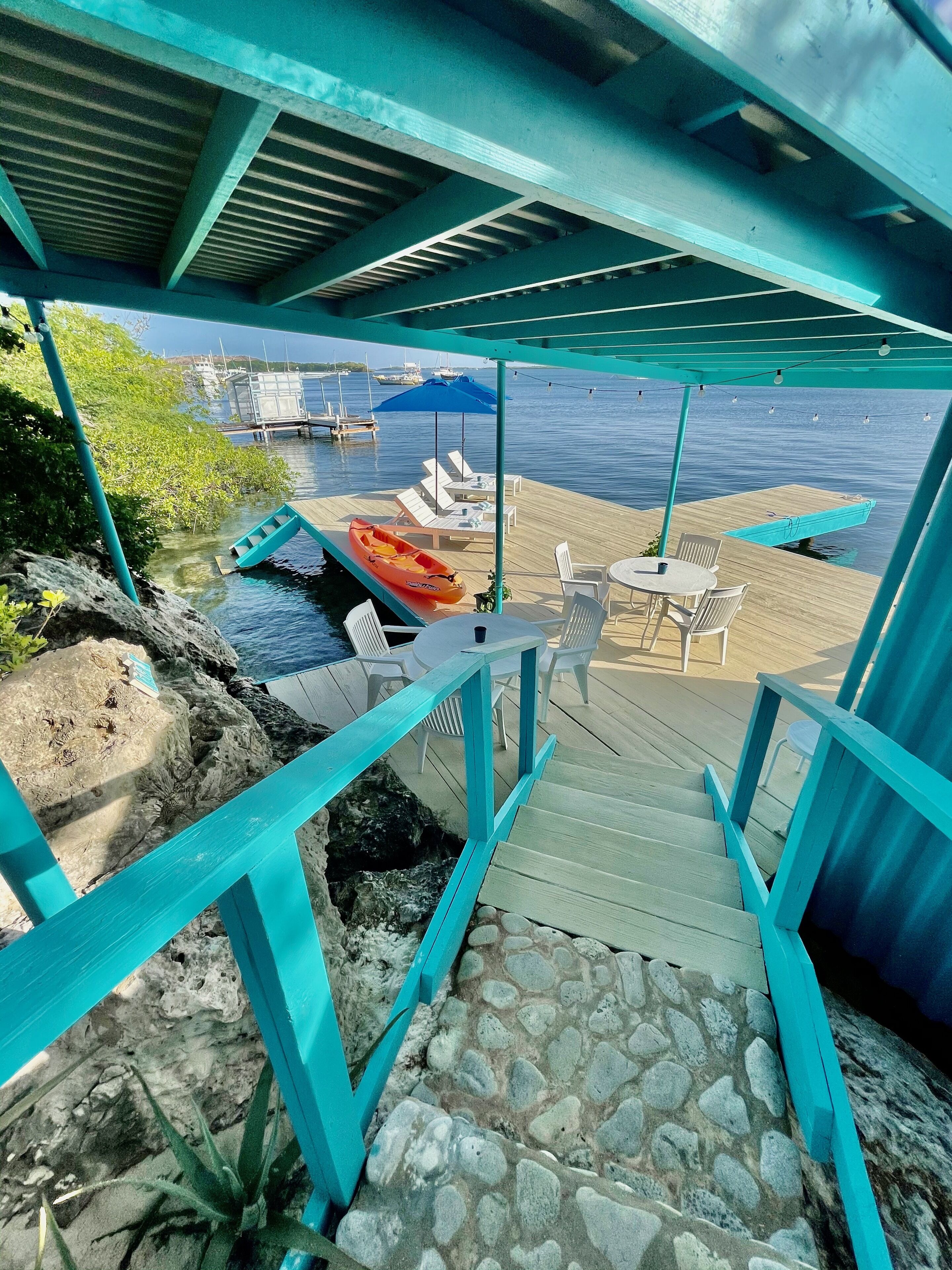 Um deck de madeira com escadaria de pedra que leva a uma área coberta para descanso, com vista para um corpo de água.
