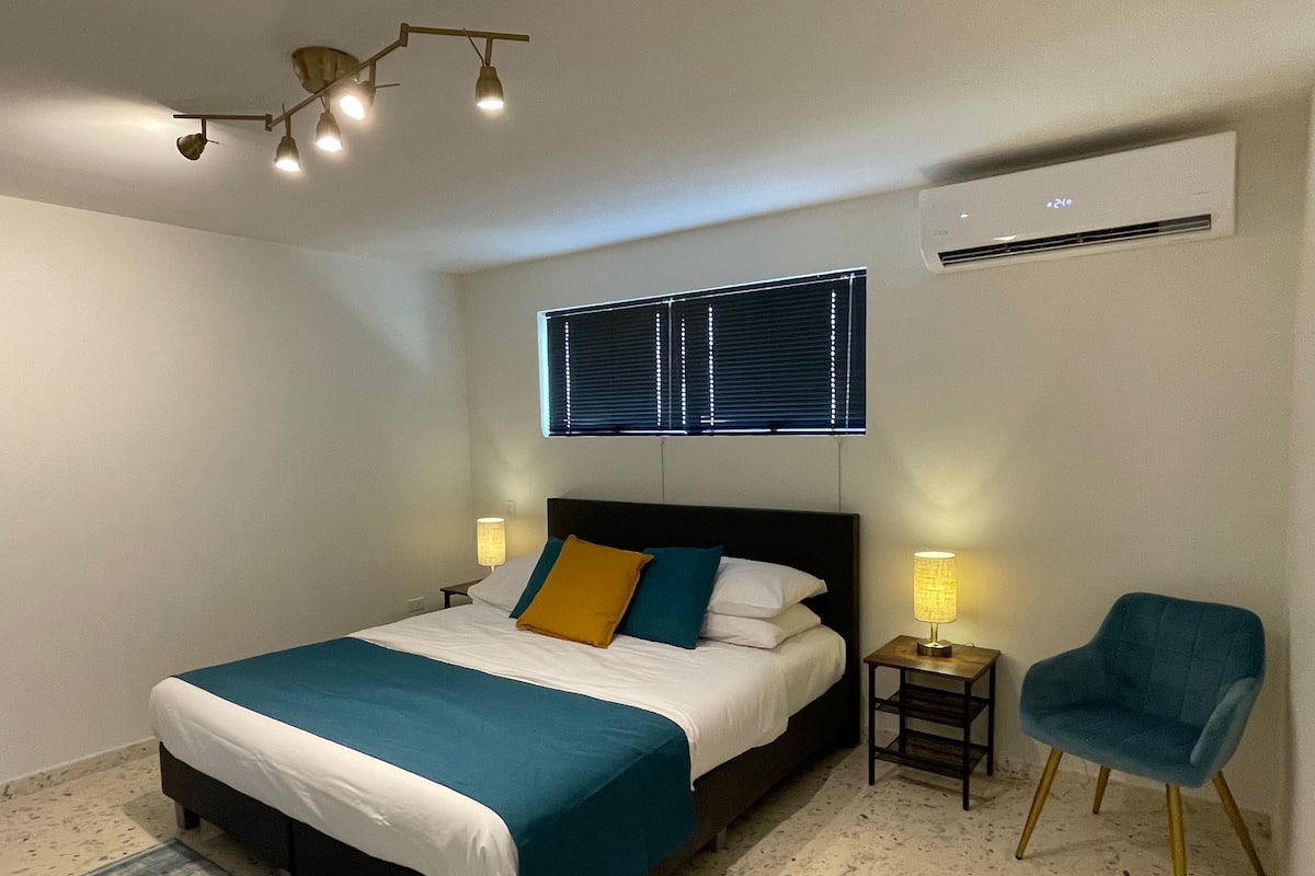 A bedroom with a bed, bedside lamps, a chair, a window with blinds, and an air conditioning unit.