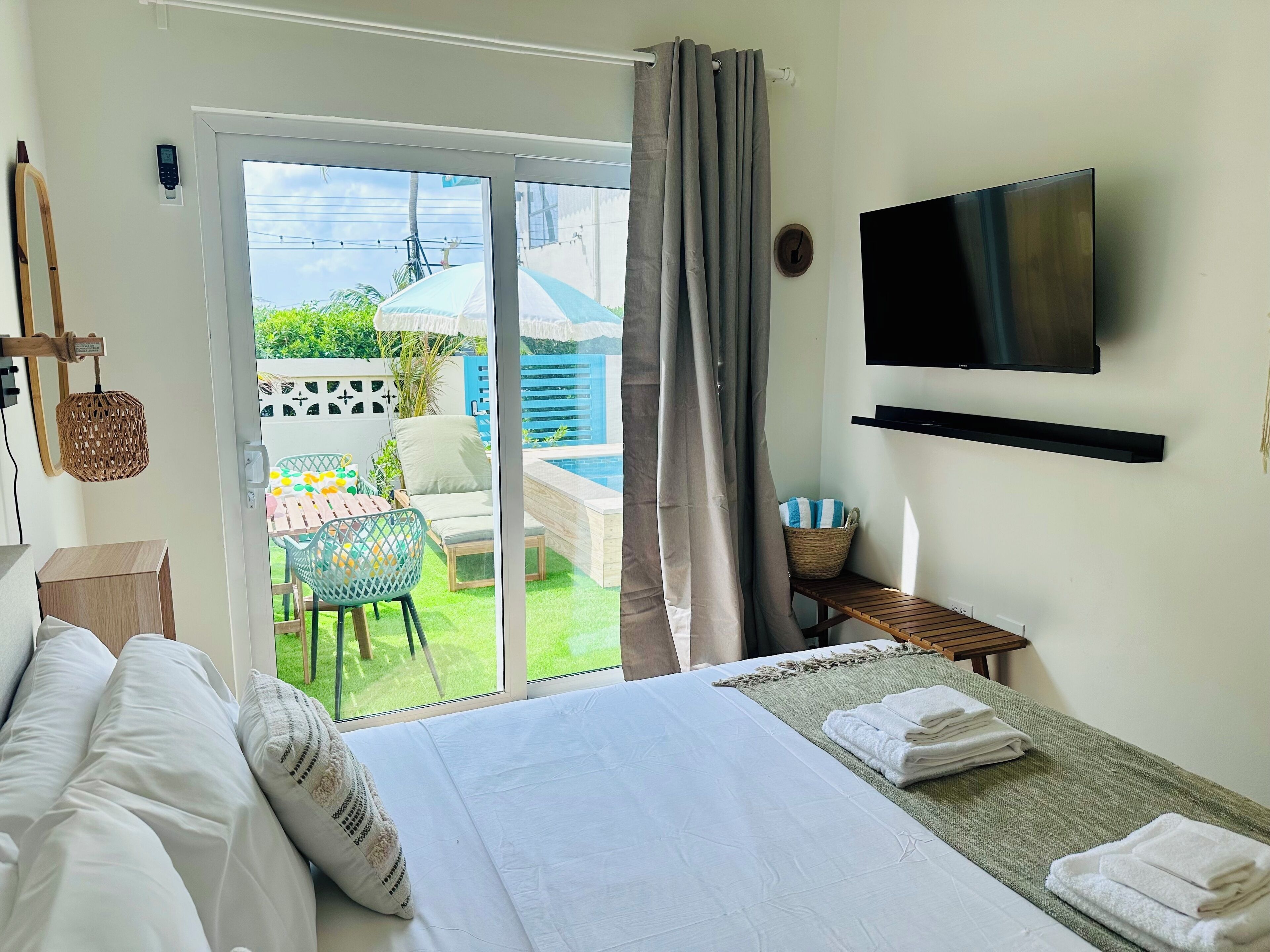 A hotel room with a bed, a TV, a TV stand, a small table with a basket, and a view of a pool and garden.