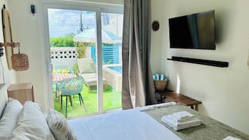 A hotel room with a bed, a TV, a TV stand, a small table with a basket, and a view of a pool and garden.