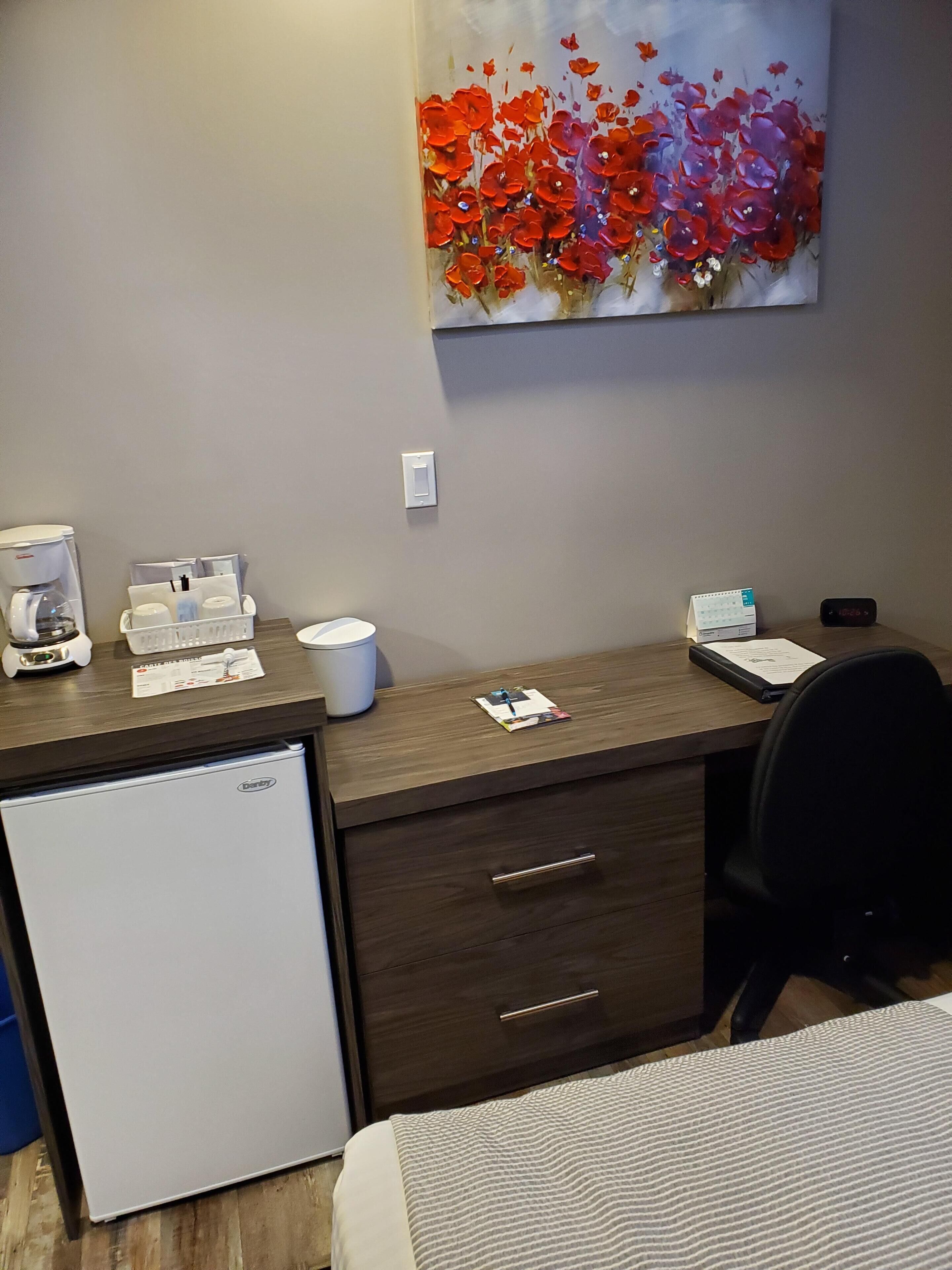 A hotel room with a small refrigerator, a desk with a coffee maker, a calendar, and a painting on the wall.