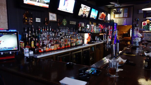 A bar with multiple screens displaying content, a well-stocked bar counter with various bottles and glasses, and a sign indicating all drinks contain alcohol. - VIP Inn and Suites (Huntsville)