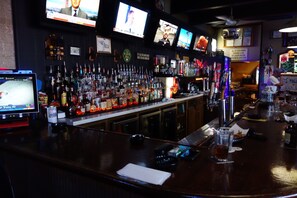 A bar with multiple screens displaying content, a well-stocked bar counter with various bottles and glasses, and a sign indicating all drinks contain alcohol. - VIP Inn and Suites (Huntsville)