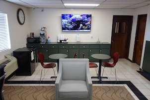 Zona de recepción con televisor, reloj, cafetera, refrigerador y dos mesas con sillas.