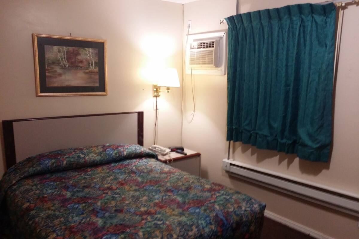 Une chambre d’hôtel équipée d’un lit, d’une lampe de chevet, d’un appareil de climatisation et d’une fenêtre avec un rideau.
