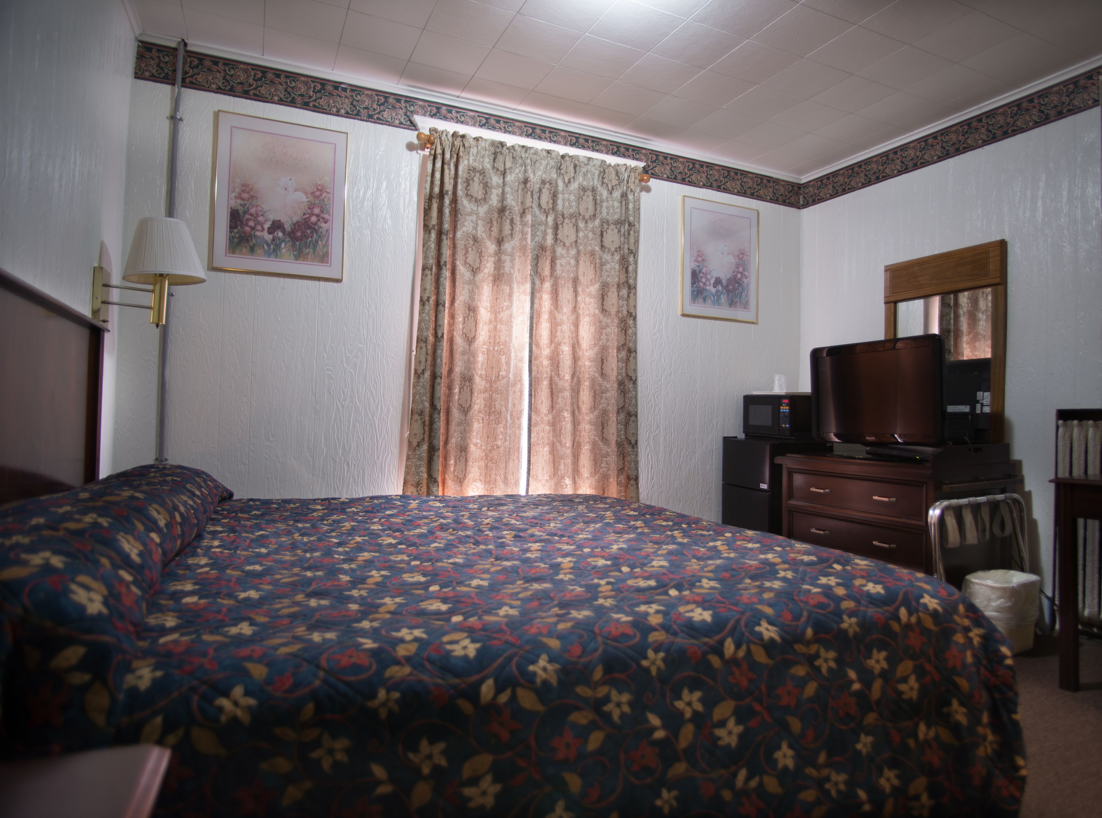 A hotel room with a large bed, a desk with a television, a lamp, and two framed pictures on the wall.
