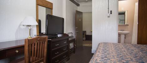 A hotel room with a bed, a desk with a television, a chair, a dresser, a mirror, and a bathroom with a sink and mirror.