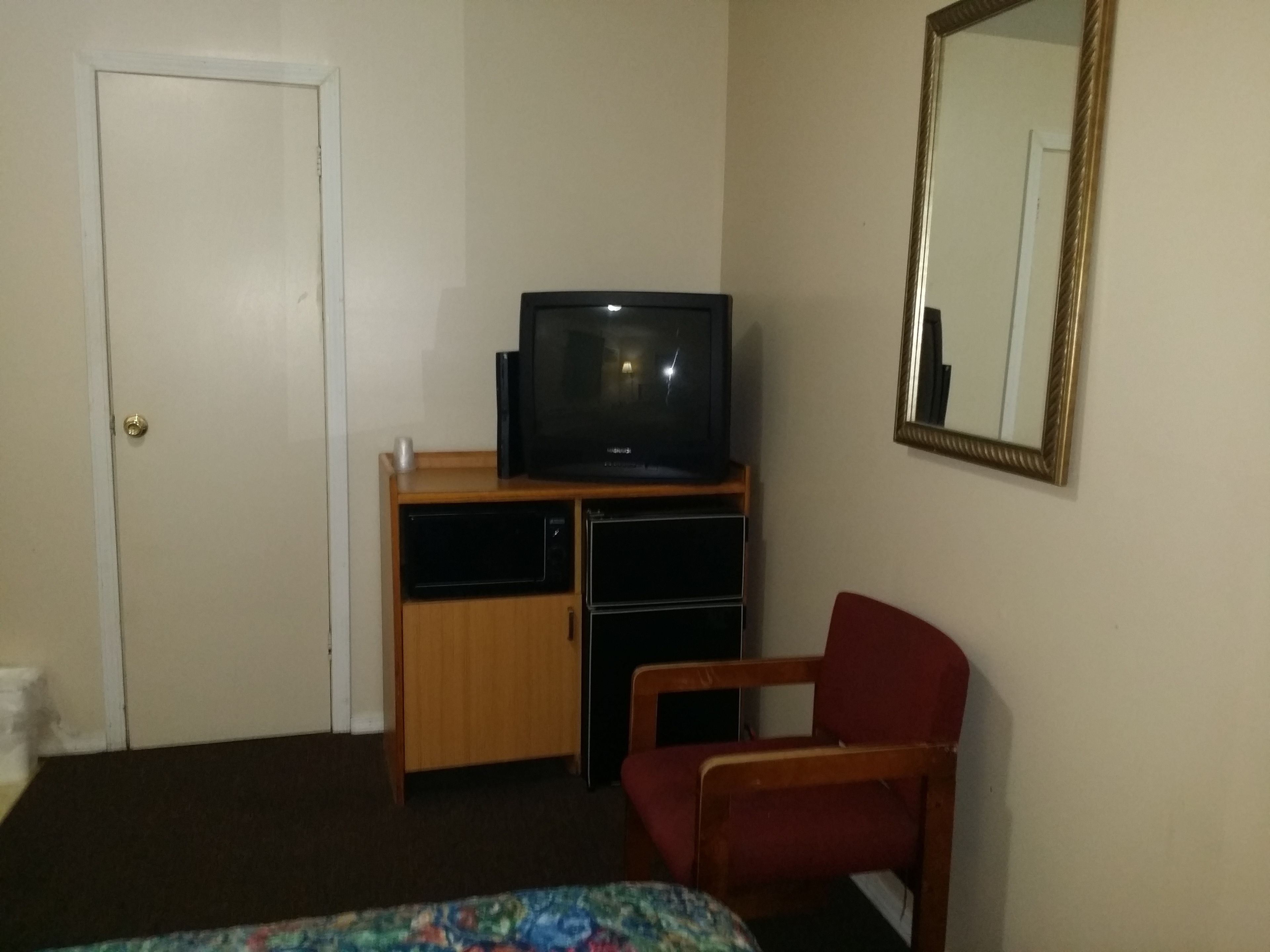 A room with a television on a wooden stand, a red chair, a bed with a floral bedspread, a door, and a mirror on the wall.