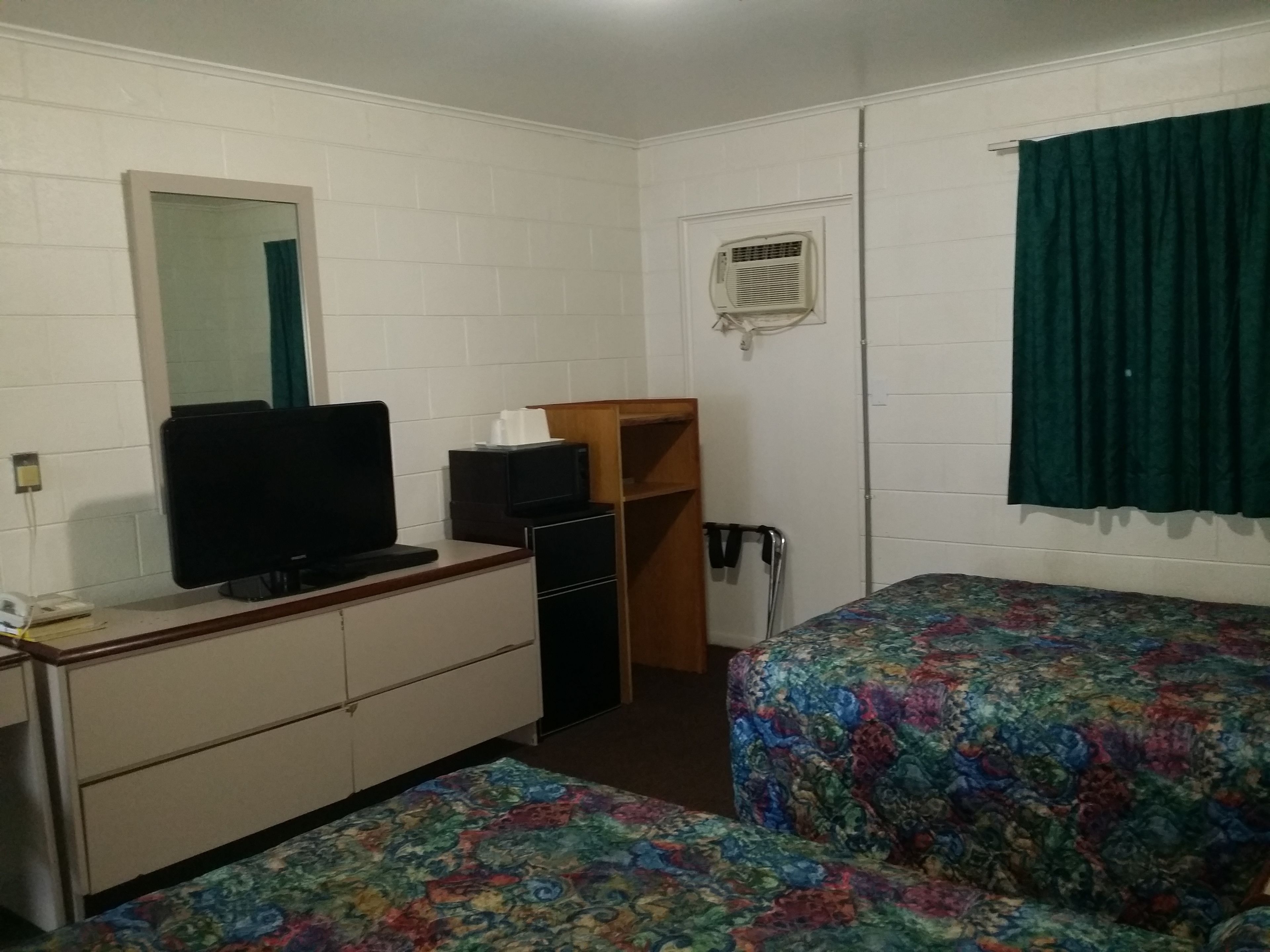 A room with a bed, a desk with a computer, a mirror, a small shelf, and a window with curtains.