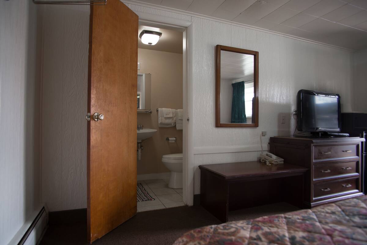 Une chambre à coucher avec une armoire en bois, une table de chevet avec un téléphone, un téléviseur posé sur une commode et un grand miroir accroché au mur.