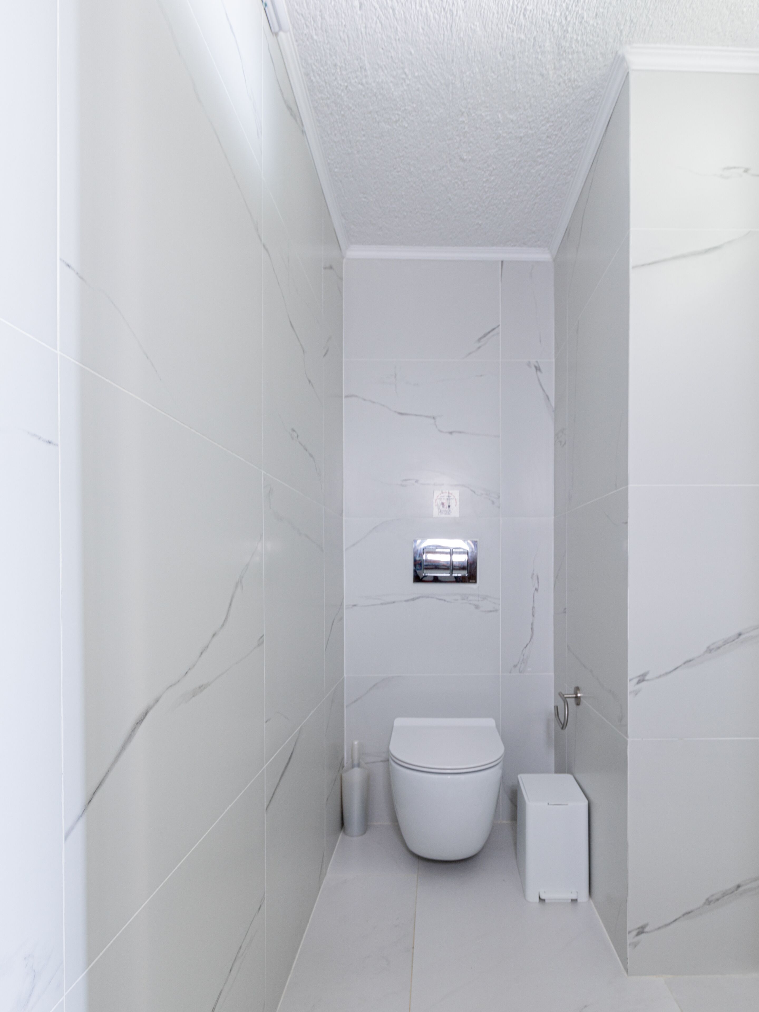 Un baño moderno con azulejos de mármol blanco, un inodoro, un bidé y un pequeño cubo de basura.