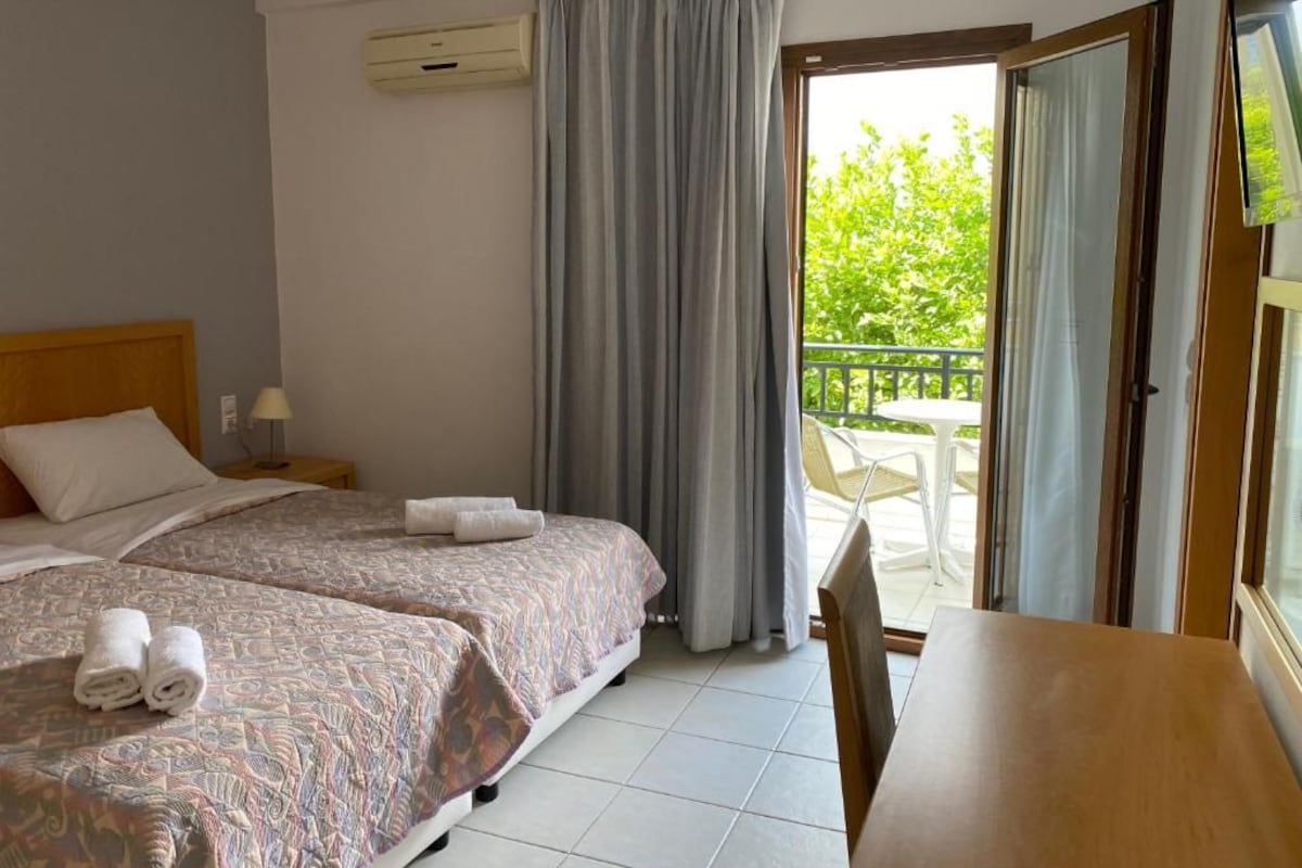 Habitación de hotel con dos camas, una mesa y balcón con vistas a zonas verdes.