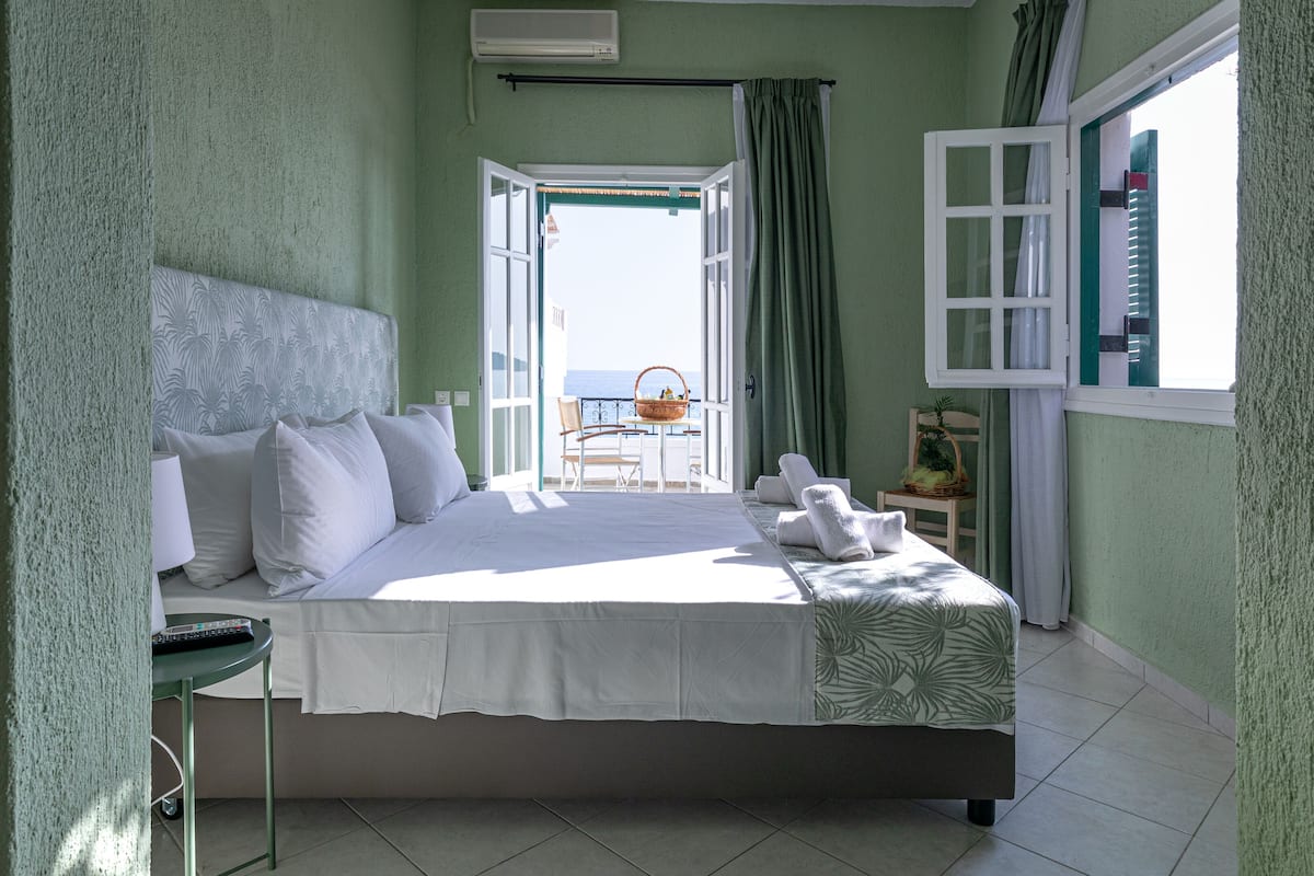A bedroom with a large bed, a view of the sea through an open window, and a balcony with a chair and table.