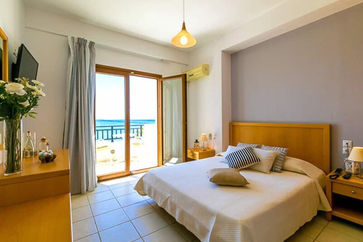 A bedroom with a large bed, a nightstand, a bedside table, a lamp, and a view of the beach through a sliding door.