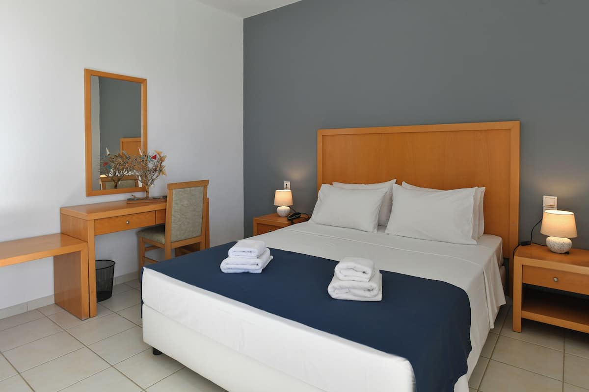 A neatly made bed with white linens and two folded towels, a wooden nightstand with a lamp, a desk with a chair, and a mirror on the wall.