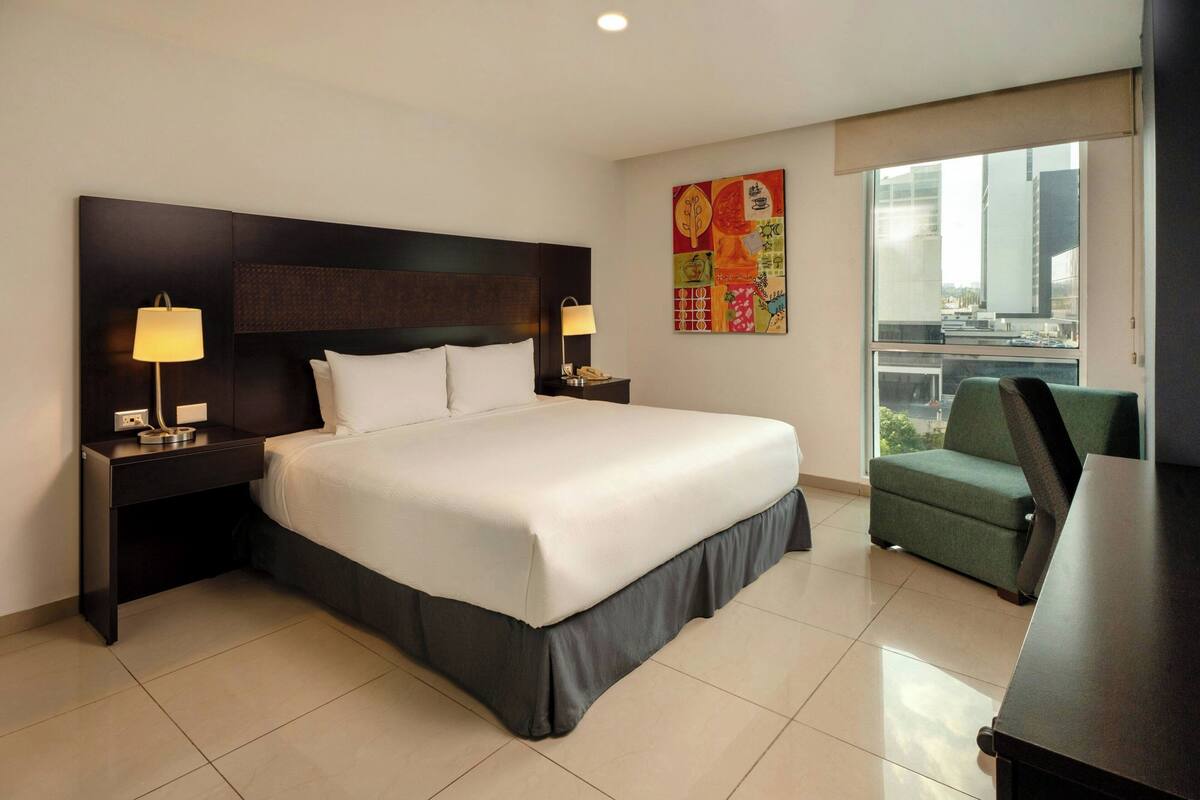 Habitación de hotel con cama, mesitas de noche, escritorio, silla, ventana con vista a la ciudad y una obra de arte colorida en la pared.