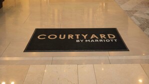 Eine Hotellobby mit einer Bank, einer Fußmatte mit dem Aufdruck 'COURTYARD BY MARRIOTT', einer dekorativen Leuchte und zwei Zimmerpflanzen.