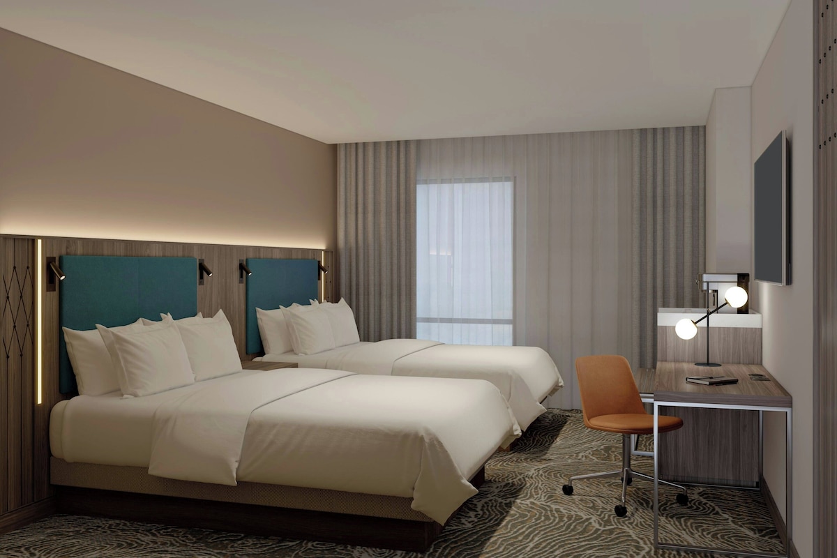A hotel room with two beds, a desk, a chair, and a TV.