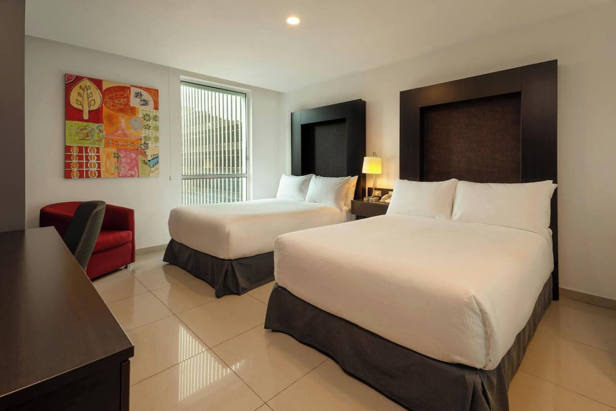 Habitación de hotel con dos camas, un escritorio, una silla, una ventana con persianas y un cuadro abstracto y colorido en la pared.