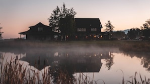 A large house with a porch and multiple windows, surrounded by trees and a body of water. - Gallatin River Lodge (Bozeman)