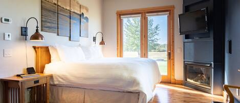 A bedroom with a large bed, wooden flooring, a fireplace, and a TV mounted on the wall.