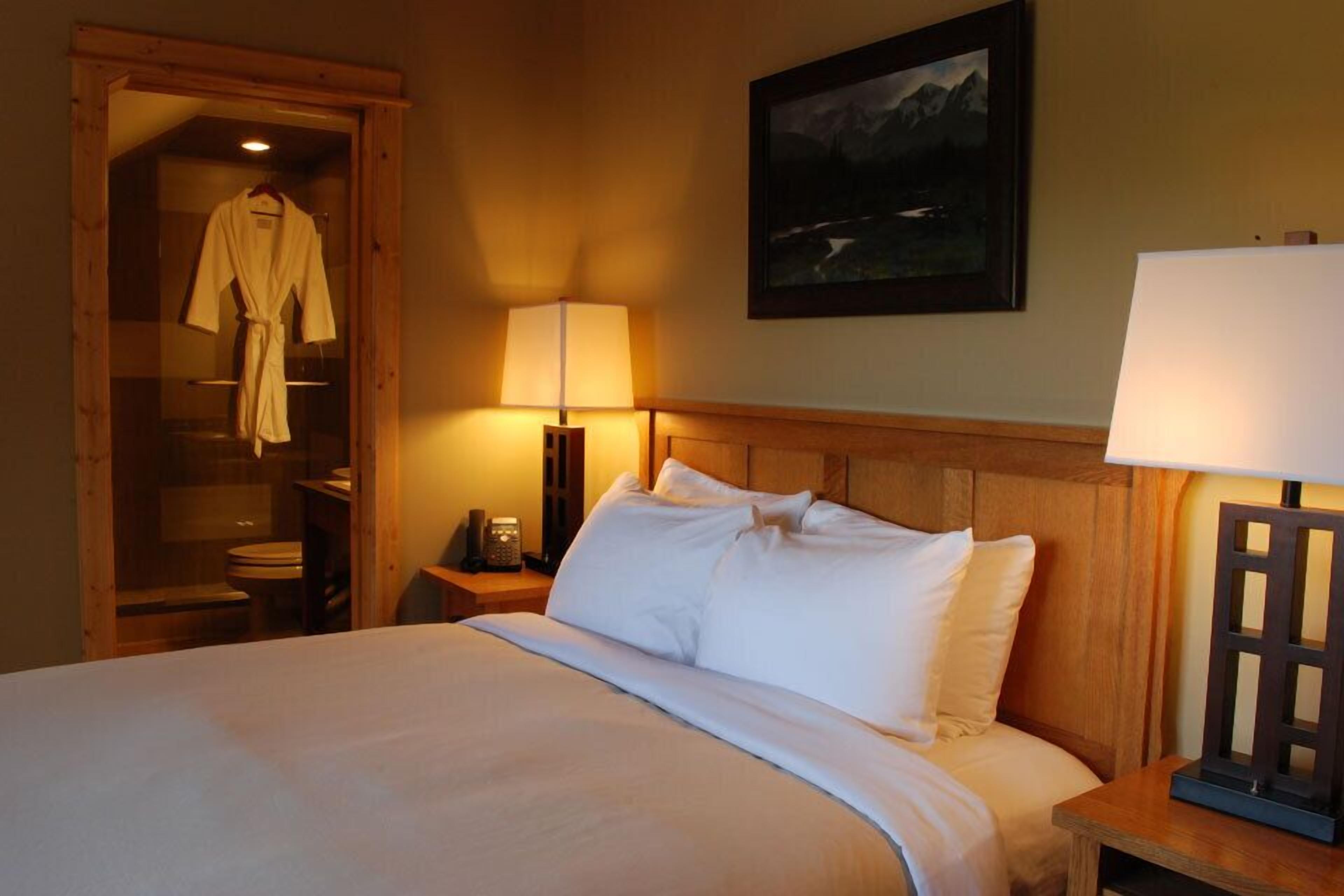 Habitación de hotel con cama, cabecera de madera, mesita de noche con lámpara, bata colgada en la puerta y un cuadro en la pared.